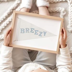 Framed pennant print with soft blue 'Breezy' lettering, held by a child while sitting on a cozy neutral rug—perfect for nursery or kids room decor.