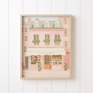 Parisian bakery-style building print in warm pastel tones, framed in rustic wood and styled on vertical paneling for a charming, French-inspired look.