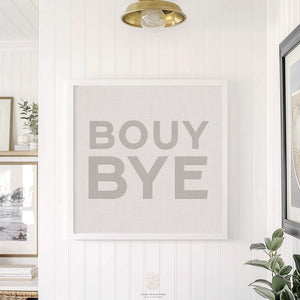 Modern neutral coastal art print reading “BOUY BYE,” framed in white and styled in a bright hallway with botanical accents.