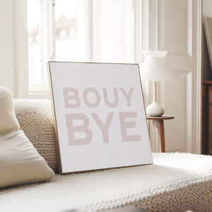 Framed blush coastal typography print reading “BOUY BYE,” styled on a textured cream blanket in a modern sunlit living room.