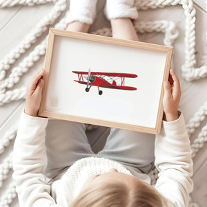 Child holding a framed red biplane art print while sitting on cozy floor with knit rug, perfect for kids' room or nursery wall decor.