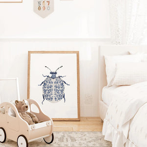 Blue and white ladybug wall art print leaning against a bedroom wall, framed in wood and styled with soft textiles and neutral nursery decor.