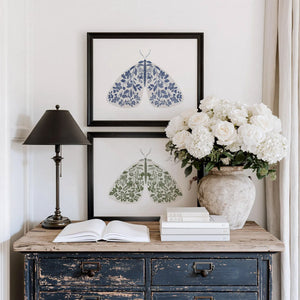 Blue floral moth print with symmetrical botanical details across its wings, displayed in a natural wood frame on a white wall.