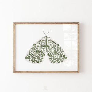 Green moth print with intricate flowers and leafy botanical details across its wings, displayed in a natural wood frame on a white wall.