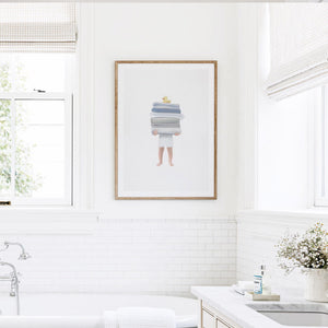 Boy Holding Towels Print featuring a whimsical illustration of a child carrying stacked towels with a rubber duck, displayed in a wood frame mockup above a white tiled bathtub.
