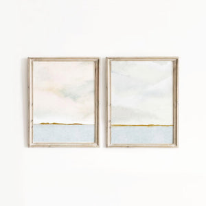 A pair of minimalist ocean horizon prints with delicate gold-green toned detailing, hung on a bedroom wall for effortless styling.