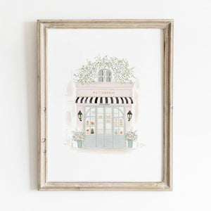 French patisserie art print with soft pastel tones and black striped awning, framed in rustic wood and styled against a white wall for elegant decor.
