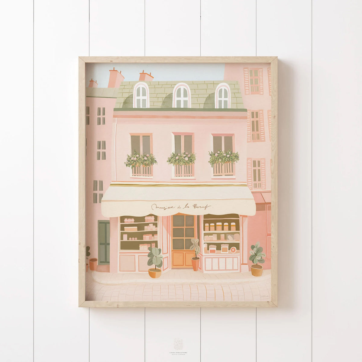 Parisian bakery-style building print in warm pastel tones, framed in rustic wood and styled on vertical paneling for a charming, French-inspired look.