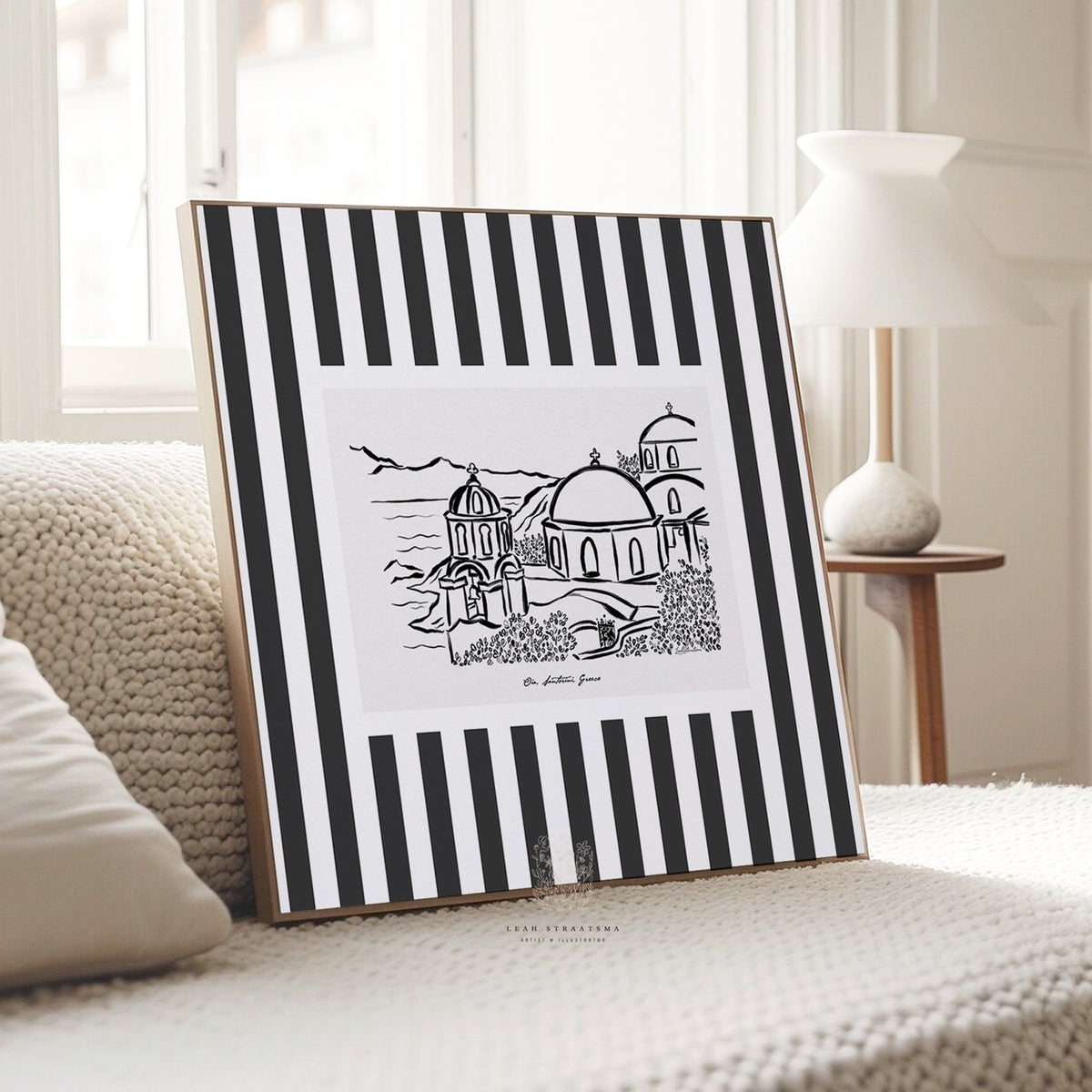 Oia Santorini printable art resting on a textured cream sofa, showing bold striped border and hand-drawn dome architecture in a cozy neutral space.