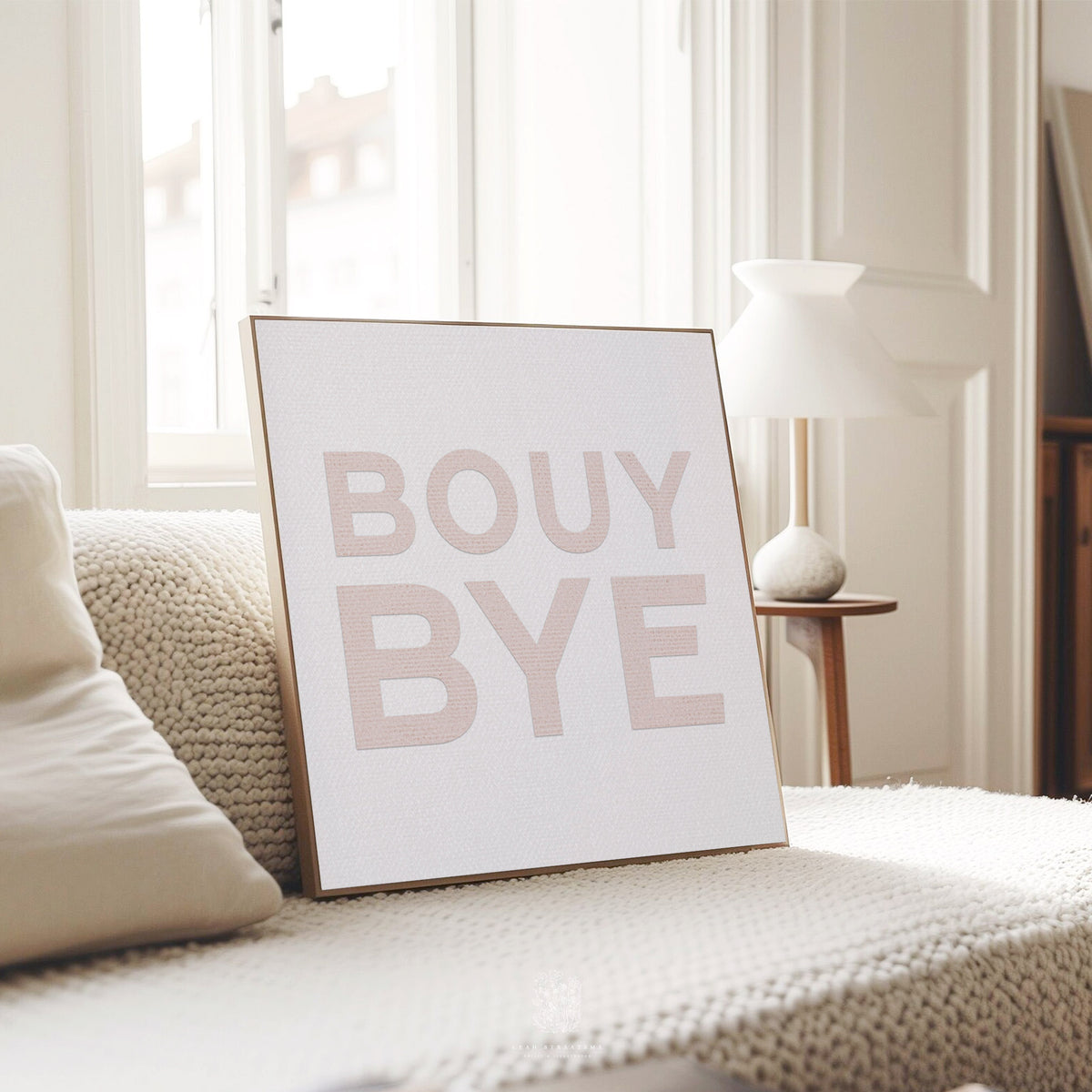Framed blush coastal typography print reading “BOUY BYE,” styled on a textured cream blanket in a modern sunlit living room.