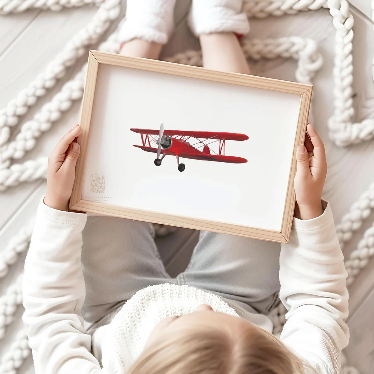Child holding a framed red biplane art print while sitting on cozy floor with knit rug, perfect for kids&#39; room or nursery wall decor.