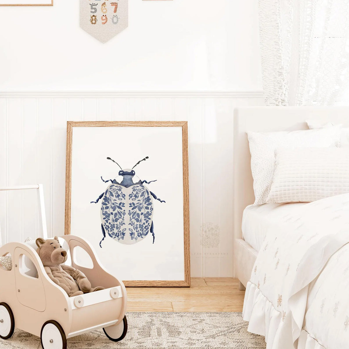 Blue and white ladybug wall art print leaning against a bedroom wall, framed in wood and styled with soft textiles and neutral nursery decor.