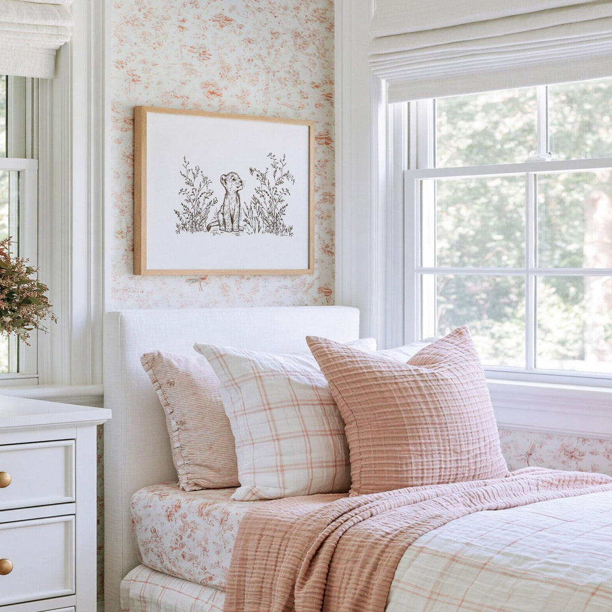 Baby lion cub print with delicate wildflowers and grasses, displayed in a natural wood frame above a bed with soft pink bedding.