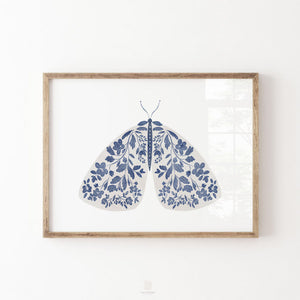 Blue floral moth print with symmetrical botanical details across its wings, displayed in a natural wood frame on a white wall.