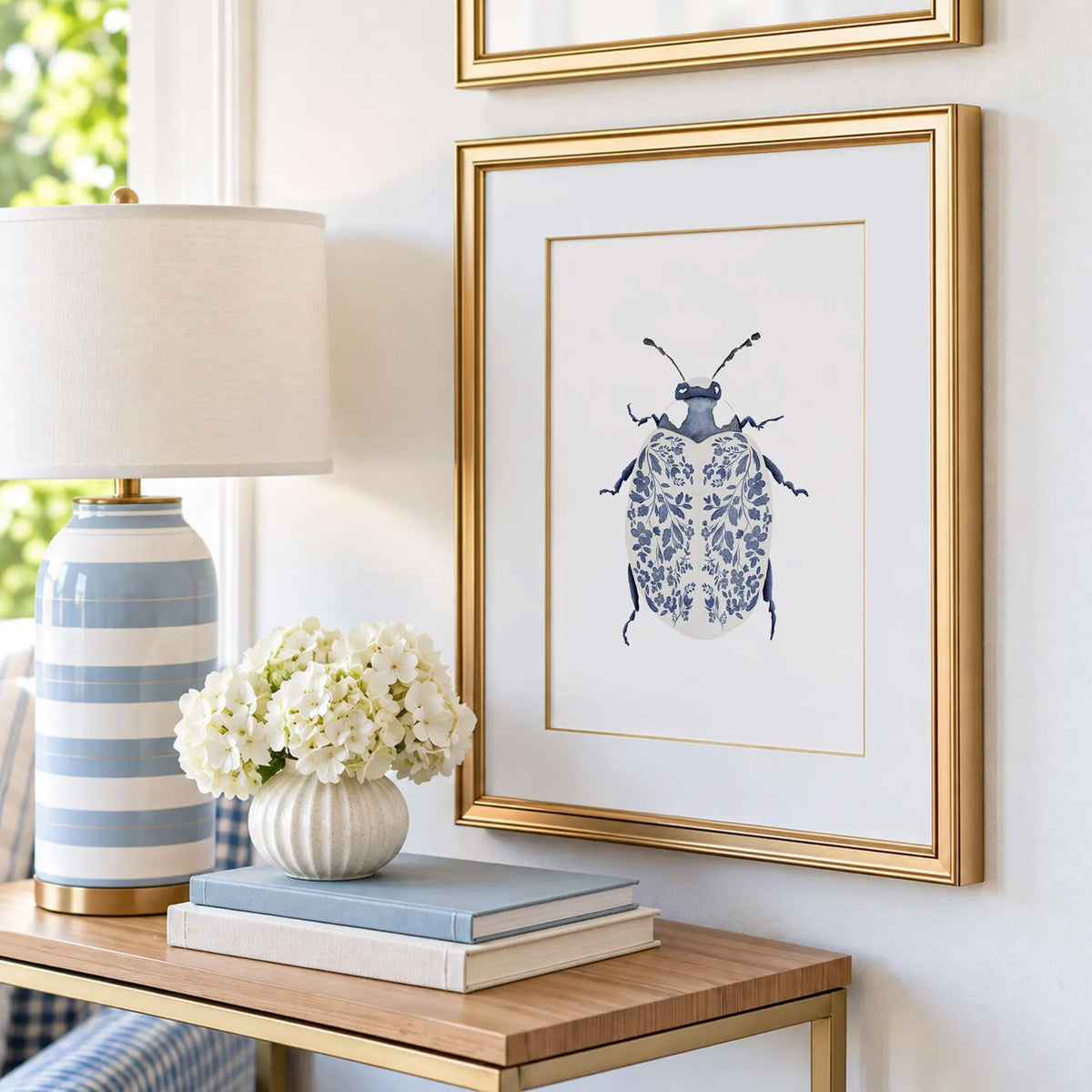 Blue Ladybug Print with navy floral-patterned wings, displayed in a gold frame beside a blue striped lamp and white flowers.