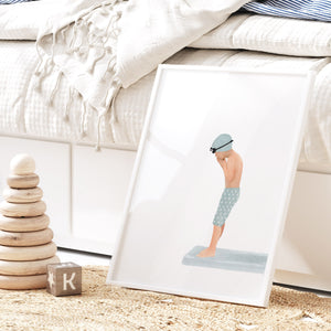 Framed print of a boy in swim trunks and goggles standing on a diving board, styled beside a bed with natural textures and cozy kids decor.