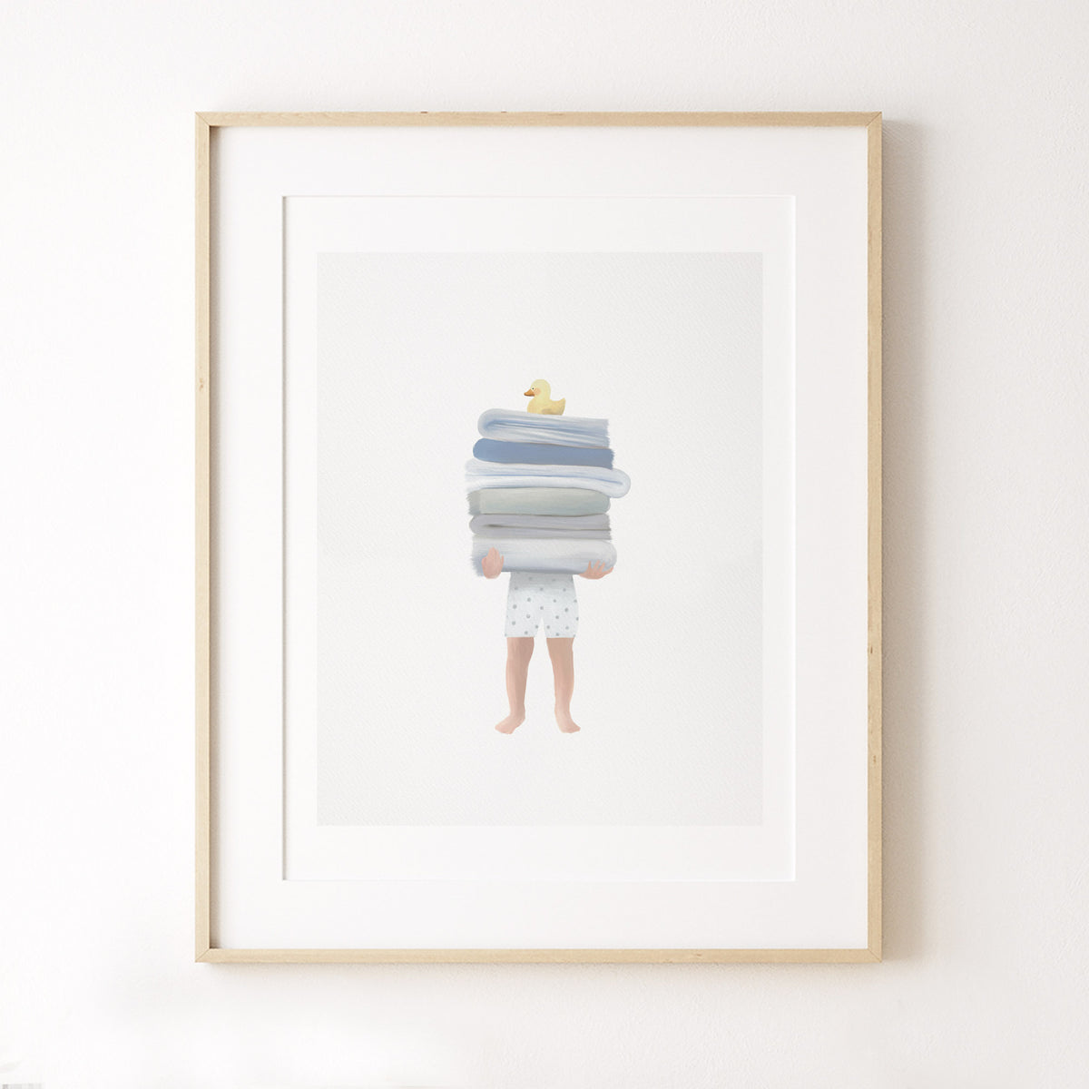 Framed print of a young boy holding folded towels with a yellow bath duck on top, styled against a white wall for kids bathroom or boys room decor.