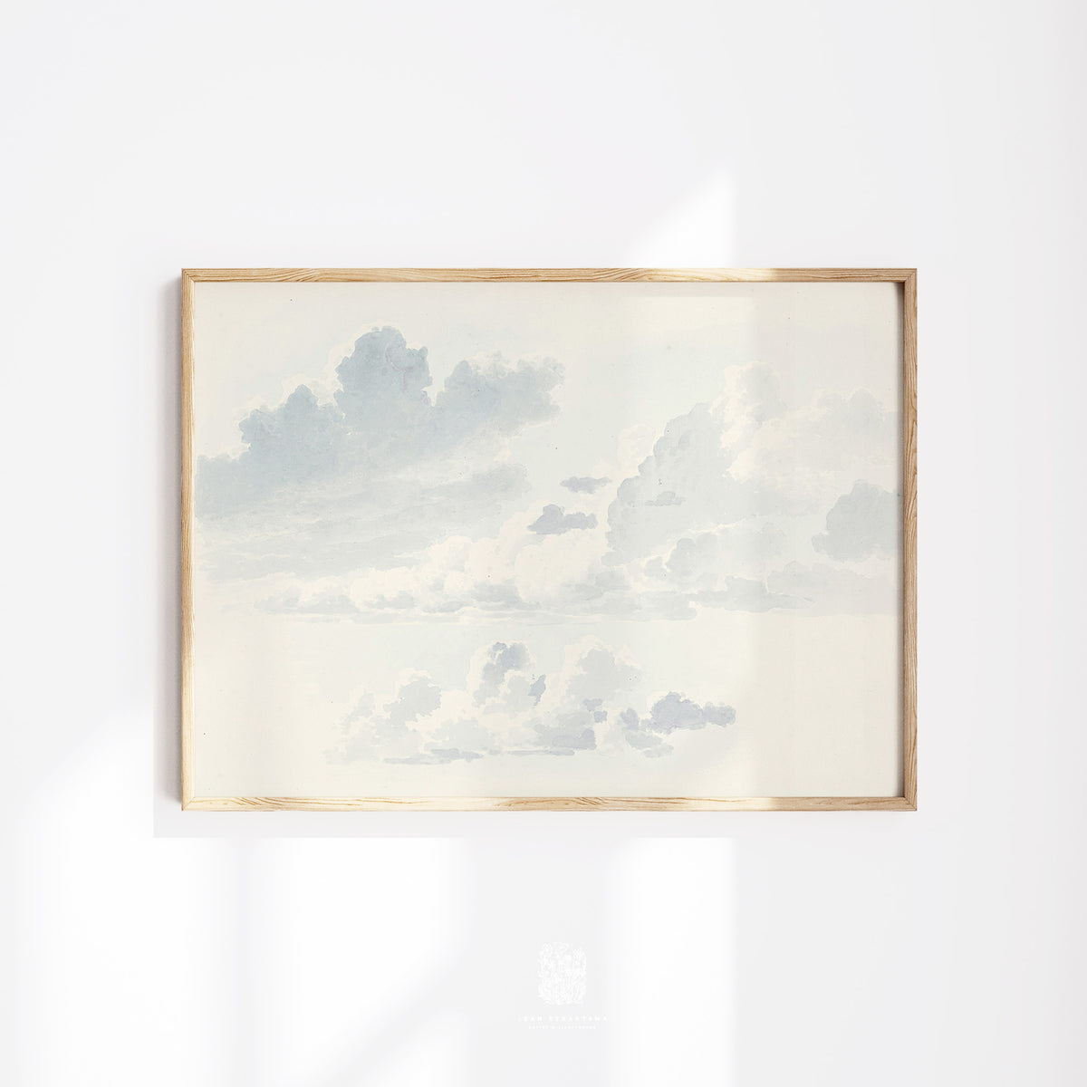 Framed cloudscape print with soft blue and cream tones, styled on a white wall with natural light and minimal golden wood frame.