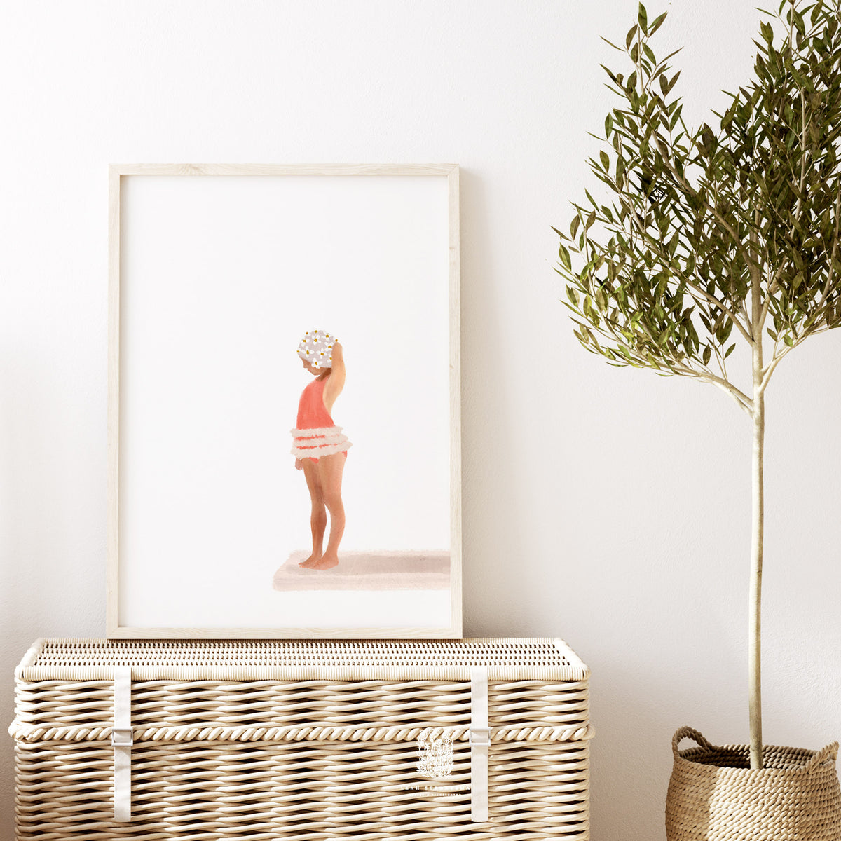Framed girl diving board print styled above a woven bench with soft white walls and natural textures, perfect for a coastal kids bathroom or nursery.