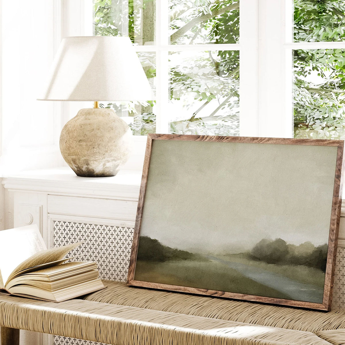 Soft green landscape art print with a misty creek scene, leaning against a window in a sunlit reading nook with natural textures.