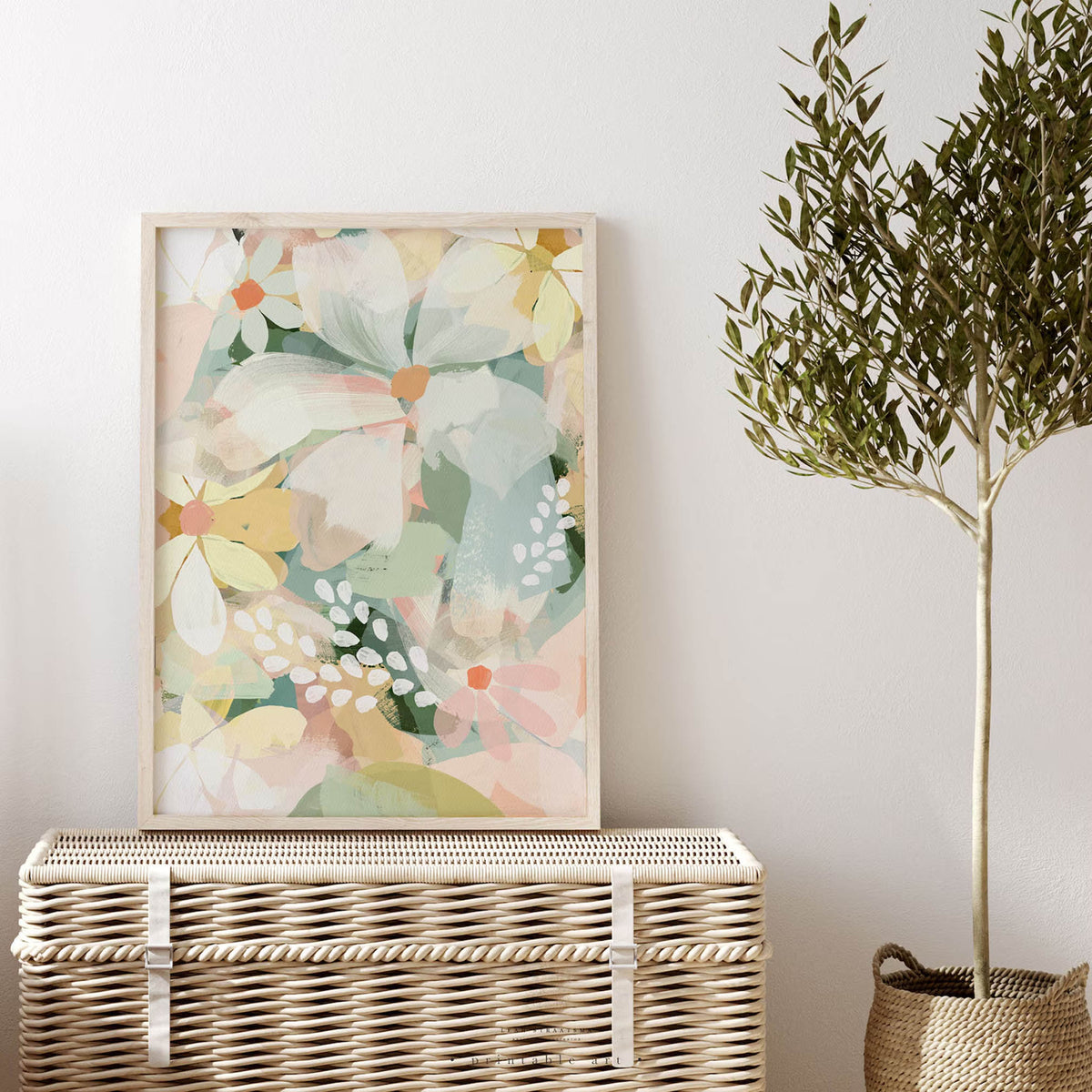 Framed abstract daisy print leaning on a rattan chest, styled with an olive tree and white wall – pastel floral wall art for modern spaces.
